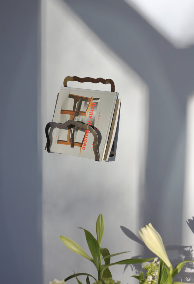 Wall-mounted magazine holder in glossy chrome with organic wave design, perfect for displaying magazines or books.